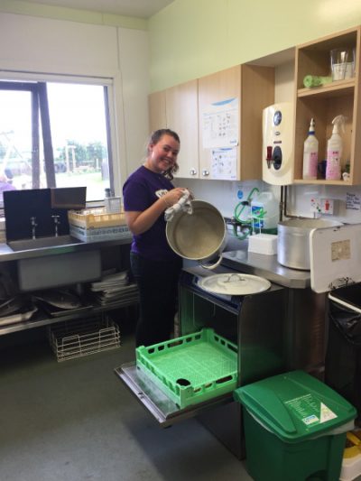 A person standing in a kitchen washing a pot and smiling at the camera.