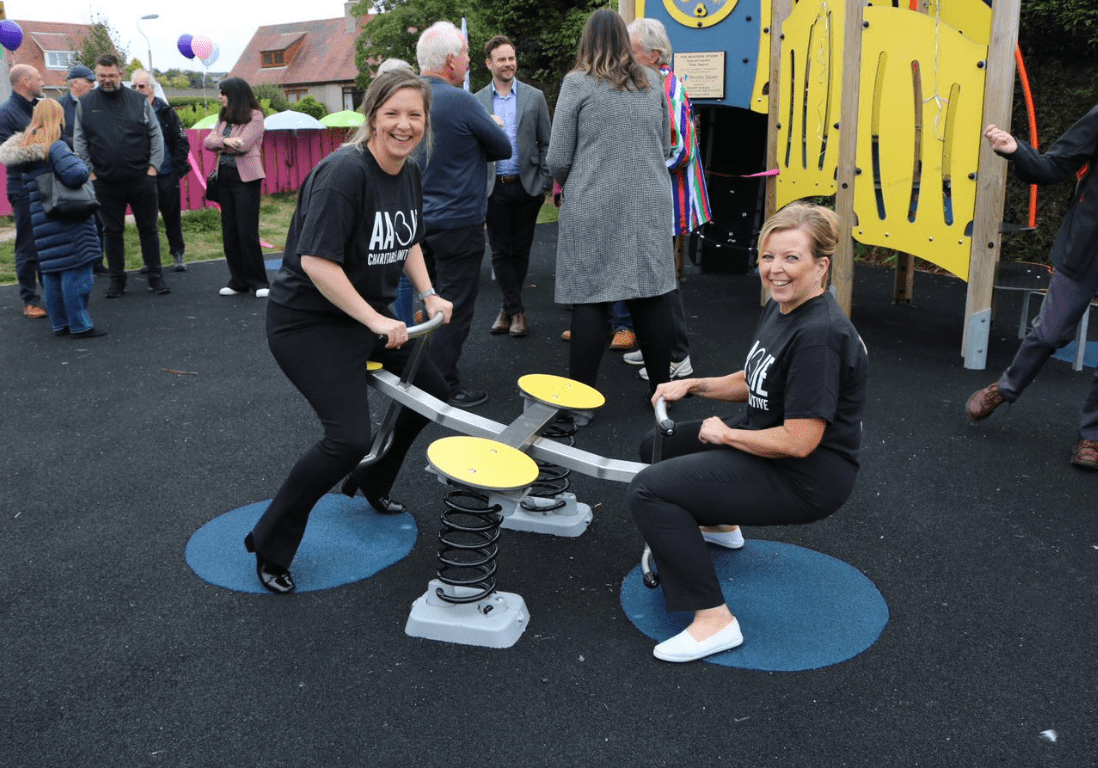 Two women on a seesaw at a playpark, one high in the air, one with feet on the ground