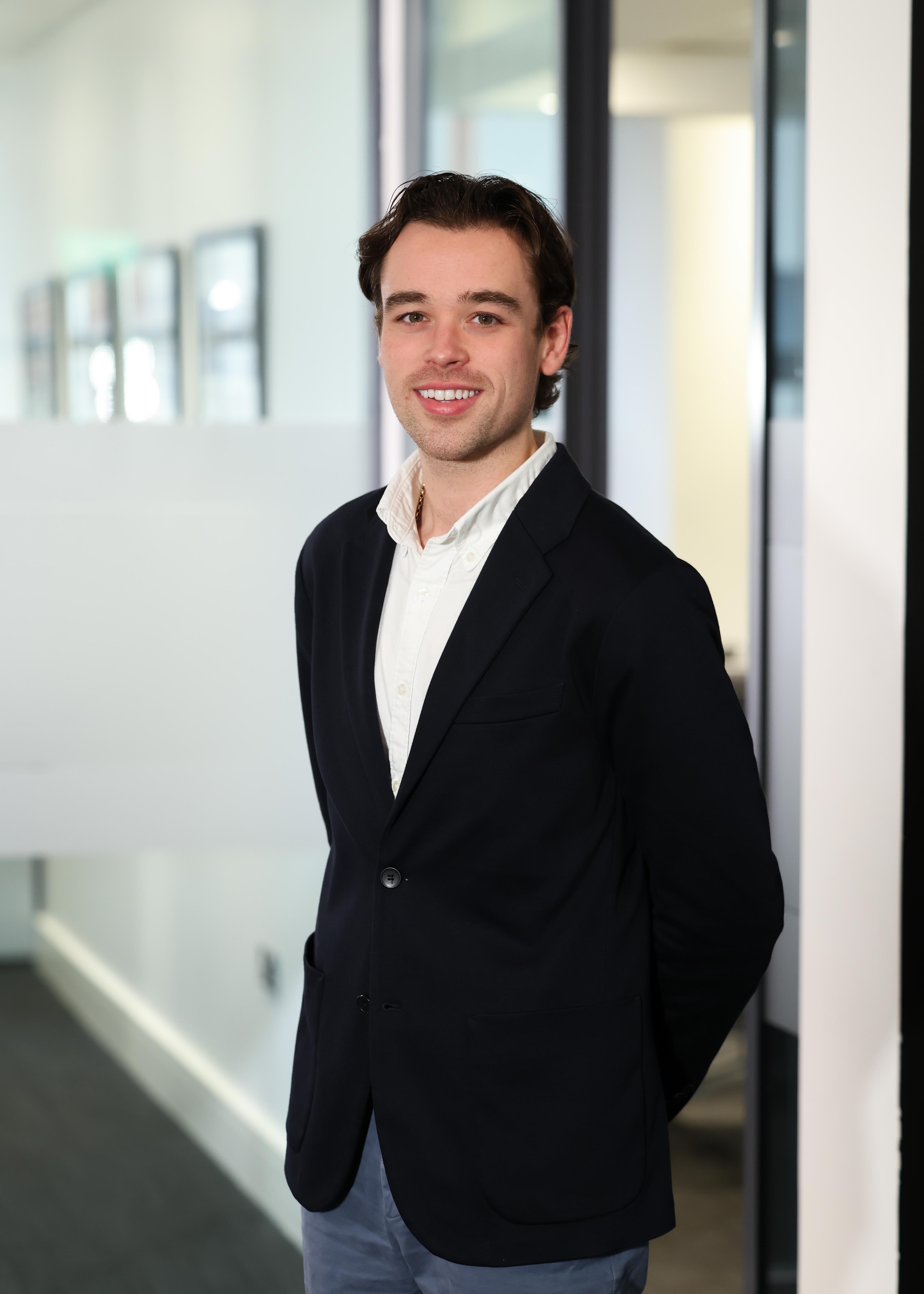 An image of Rory MaCallister wearing a black suit jacket with a white shirt. He is standing in the office.