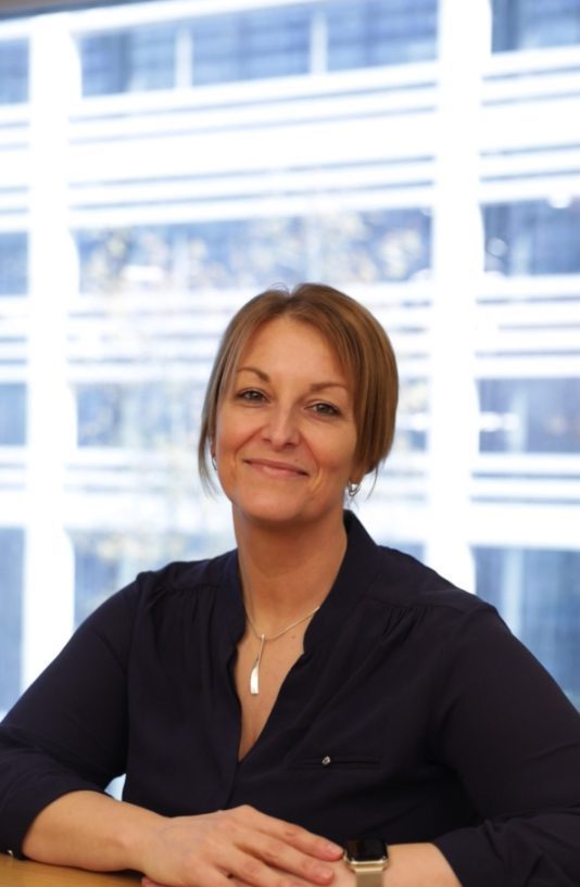 An image of Rachel Coupland smiling at the camera and sitting at her desk.