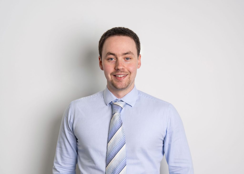 An image of Fraser White wearing a blue shirt and tie. He is smiling at the camera.