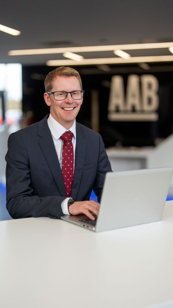 An image of Andrew Wester sitting at a desk working on his laptop. He is wearing a grey suit jacket with a burgandy tie.