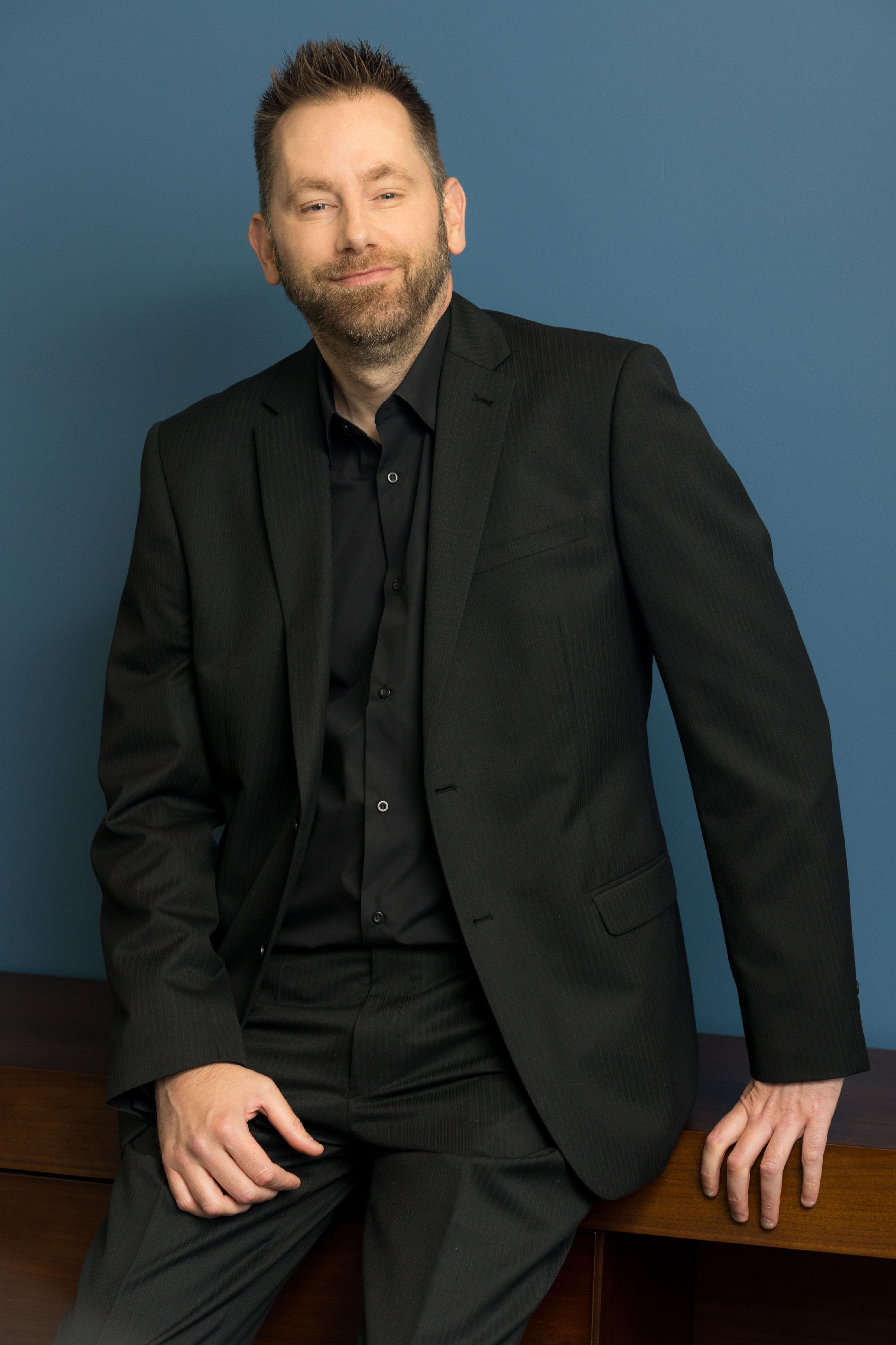 An image of Damien Coughlan wearing a black blazer with a black shirt. He is resting on a desk and smiling at the camera.
