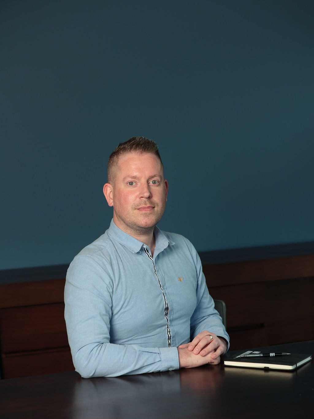 An image of David Felton wearing a blue shirt and smiling at the camera. He is sitting down with his hands on the desk.
