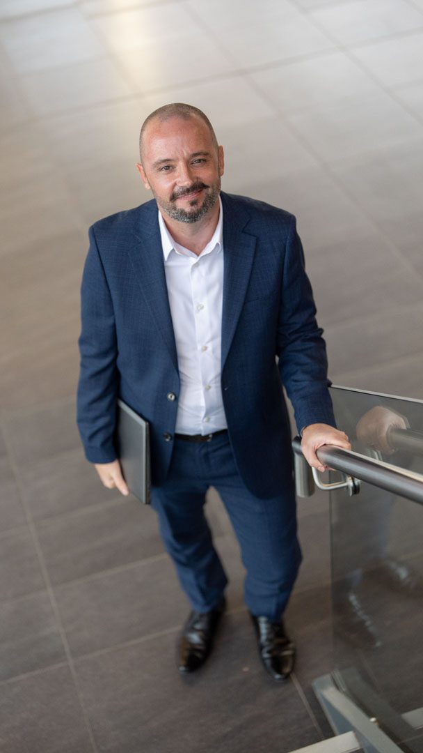 An image of Greg McSneddon wearing a suit with a white shirt. He is standing at the bottom of the stairs.