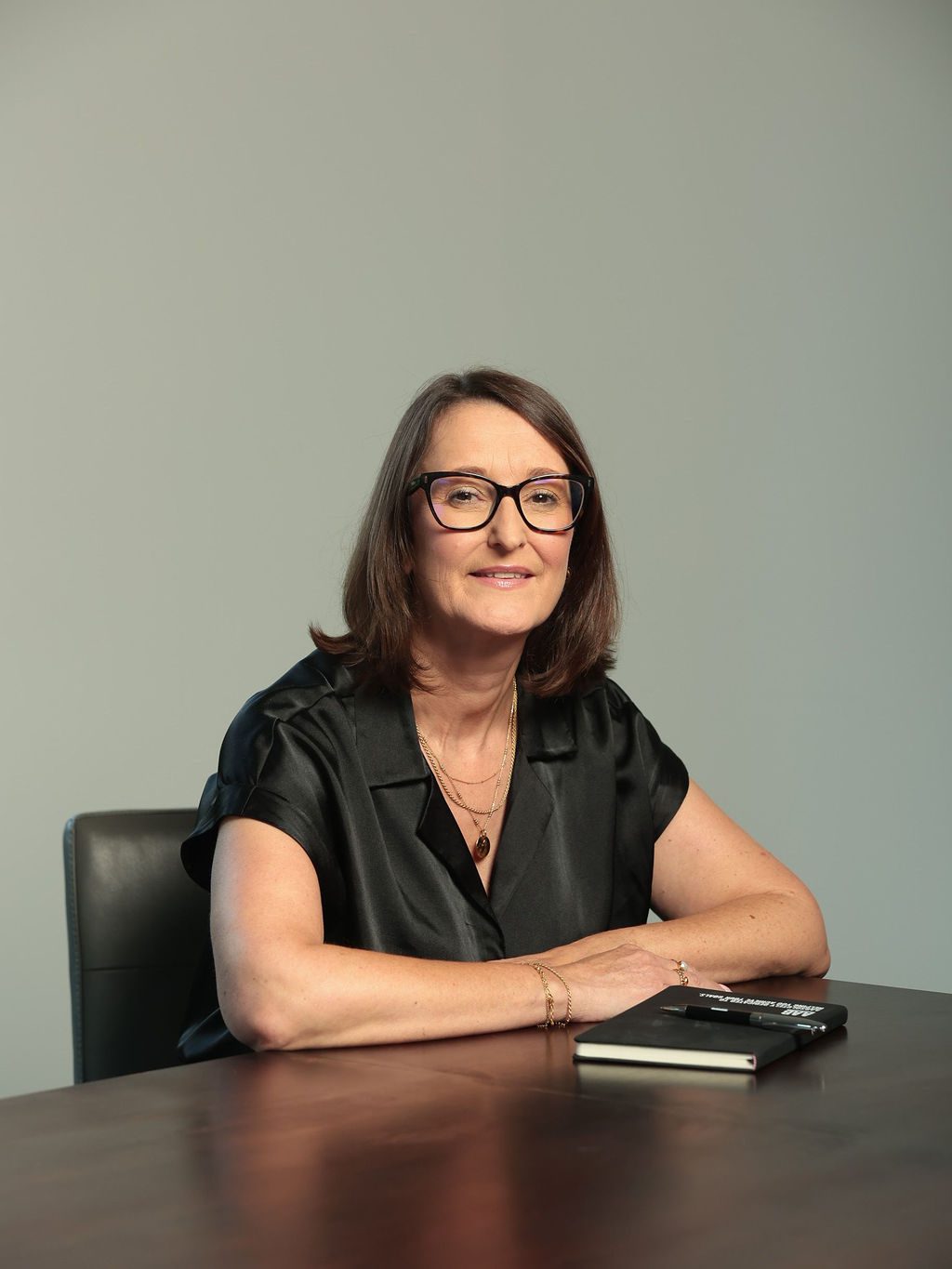 An image of Francesca Powell sitting at a desk with her AAB Wealth notebook and smiling at the camera. She is wearing a black top and black glasses.