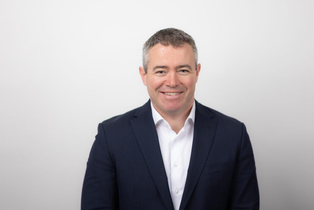 An image of Colm Duggan wearing a black suit and smiling at the camera.