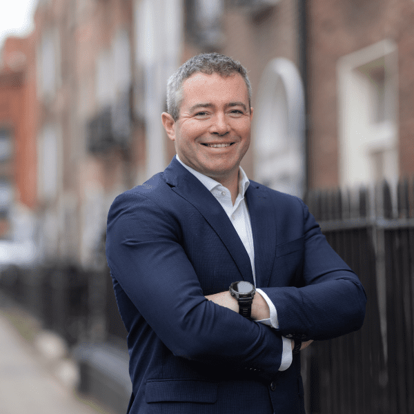 An image of Colm Duggan wearing a suit and tie. He is standing outside with his arms crossed.