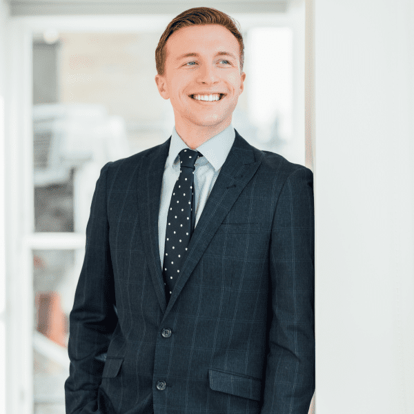 An image of Fraser Mackay wearing a black suit with a tie. He is standing in the office looking out the window.