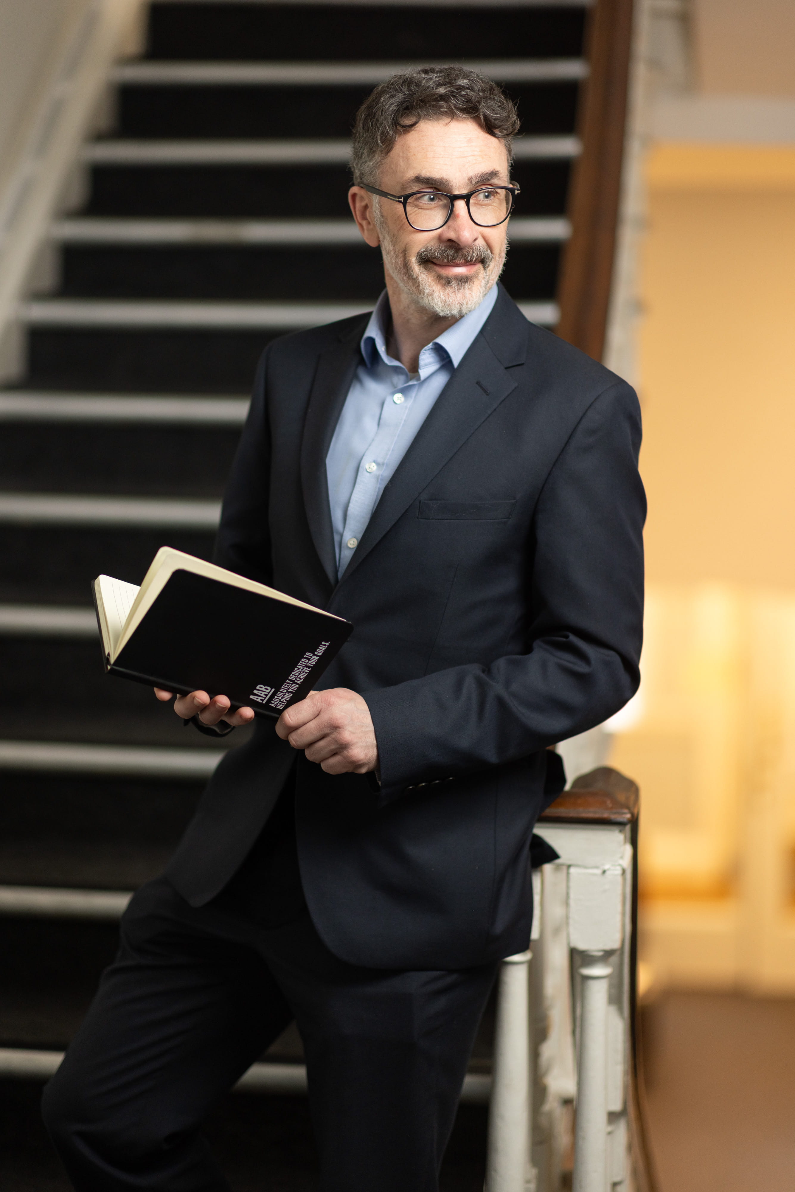 An image of Darren Benham wearing a black suit jacket and blue shirt. He is standing on the staircase, holding an AAB notebook.